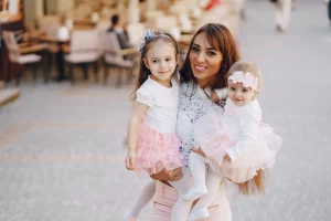 Mujer posa sonriente con 2 niñas en brazos, las niñas visten tutús de ballet rosados y están en mitad de una calle cerca de un restaurante de día