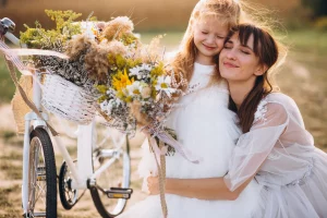 Madre que se va a casar posa con los ojos cerrados abrazando a su hija que también cierra los ojos junto a un campo y al lado de una bicicleta blanca con una cesta de flores amarillas, blancas y verdes